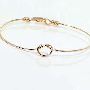 May include: A gold-toned bangle bracelet with a knot design. The bracelet is made of thin wire and has a simple, elegant look.