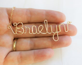 May include: Gold wire necklace with the name "Braelyn" spelled out in cursive.