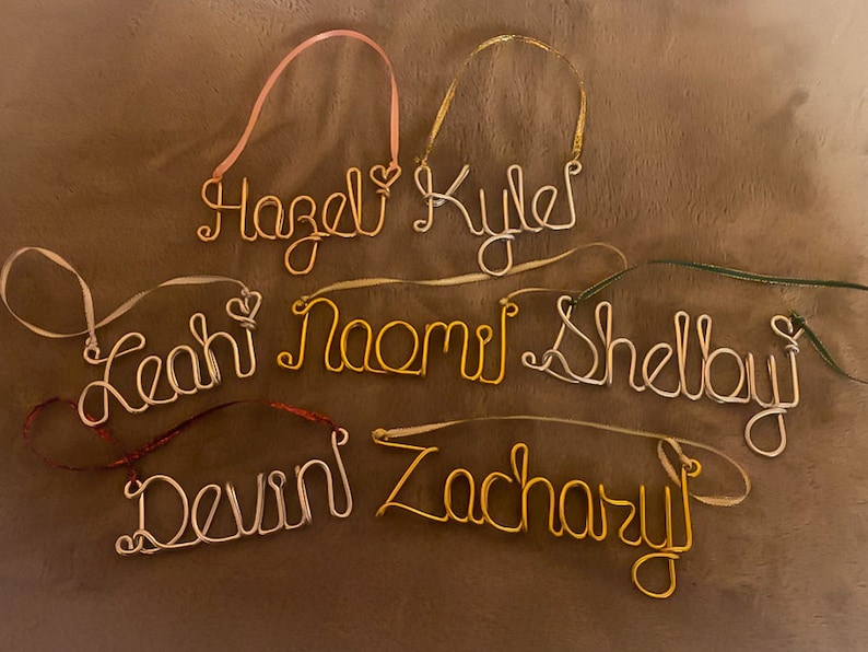 May include: Handmade wire name ornaments in gold and silver tones, each with a ribbon for hanging. Names include Hazel, Kyle, Leah, Naomi, Shelby, Devin, and Zachary. A personalized gift idea for Christmas or other special occasions.