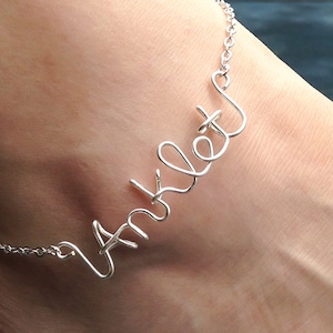 May include: A silver-colored wire anklet spelling out the word "Anklet" in cursive script. The jewelry is worn around the ankle and attached with a delicate chain.