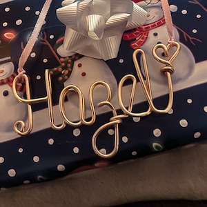 May include: A gold wire name tag spelling "Hazel" with a heart-shaped loop on the "J". The tag hangs from a pink ribbon, resting on a gift wrapped in blue paper with white polka dots and snowman illustrations.