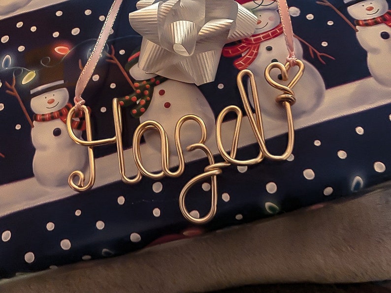 May include: A gold wire name tag spelling "Hazel" with a heart-shaped loop on the "J". The tag hangs from a pink ribbon, resting on a gift wrapped in blue paper with white polka dots and snowman illustrations.