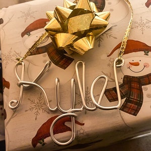 May include: A silver wire ornament spelling "Riley" is placed on a gift wrapped in snowman-themed paper. A large gold bow sits atop the gift, with a gold ribbon attached to the ornament.