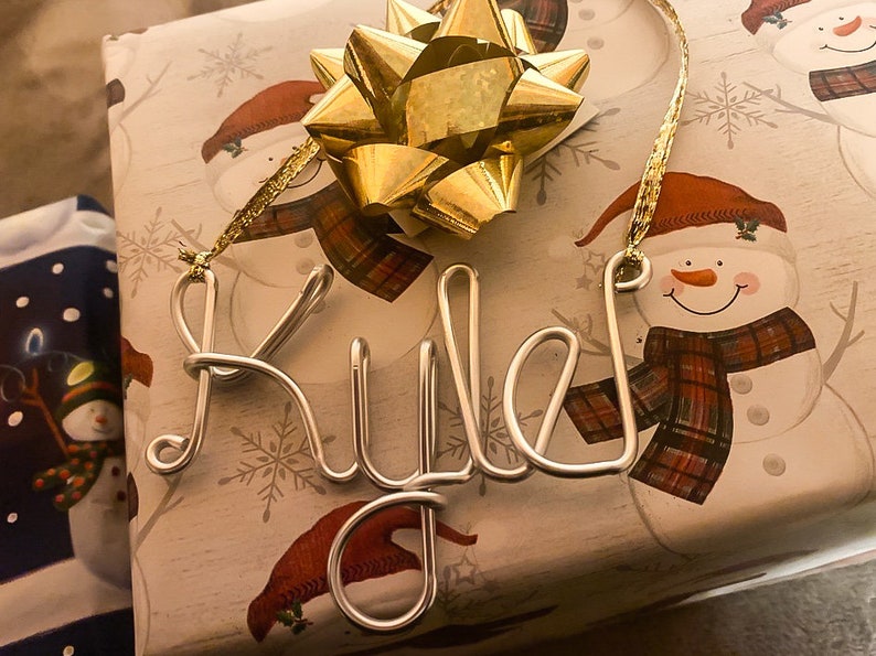 May include: A silver wire ornament spelling "Riley" is placed on a gift wrapped in snowman-themed paper. A large gold bow sits atop the gift, with a gold ribbon attached to the ornament.