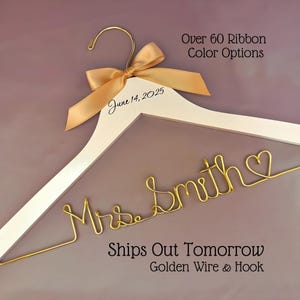 May include: A white wooden clothes hanger featuring a gold metal hook and a gold satin ribbon bow. The hanger is personalized with the text "June 14, 2025" and gold wire shaped into the name "Mrs. Smith" ending with a small heart symbol.