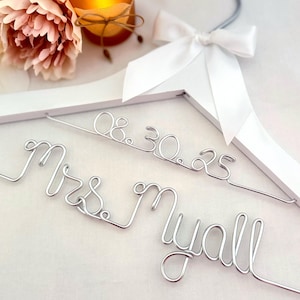 May include: A white wooden clothes hanger featuring a silver wire script that reads Mrs. Myall and the date 08.30.25. The hanger is adorned with a white satin ribbon bow tied at the top near the silver metal hook, creating a decorative display.