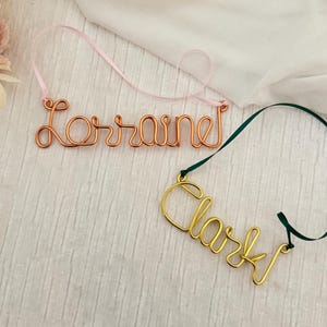 May include: Two wire name ornaments. One is copper-colored and spells "Lorraine" with a pink ribbon. The other is gold-colored and spells "Clark" with a green ribbon. Both are on a white background.