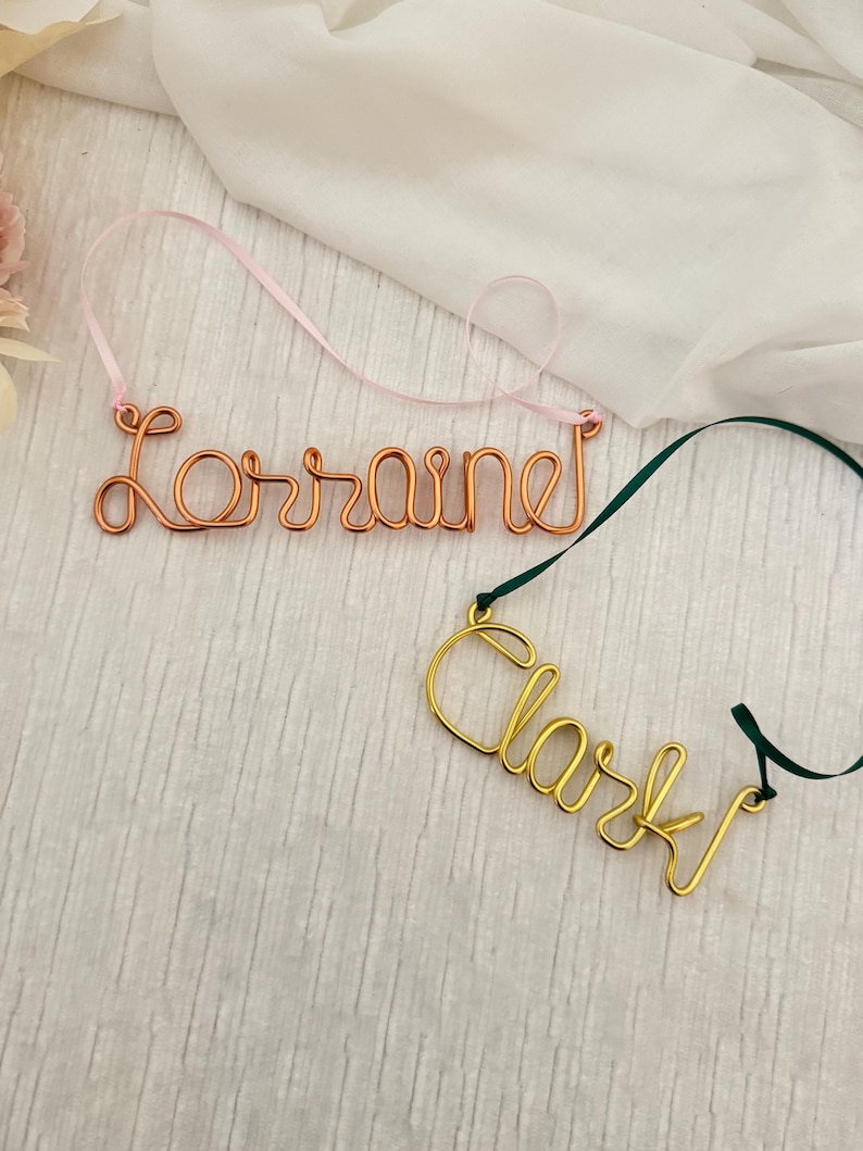 May include: Two wire name ornaments. One is copper-colored and spells "Lorraine" with a pink ribbon. The other is gold-colored and spells "Clark" with a green ribbon. Both are on a white background.