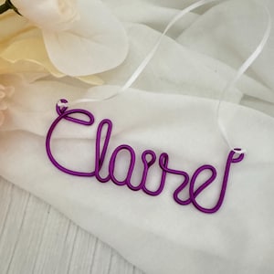May include: A purple wire name necklace spelling "Claire" in cursive script. The necklace is displayed on a white fabric with a white ribbon attached for hanging. A white flower is in the background.
