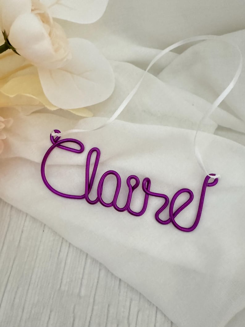 May include: A purple wire name necklace spelling "Claire" in cursive script. The necklace is displayed on a white fabric with a white ribbon attached for hanging. A white flower is in the background.