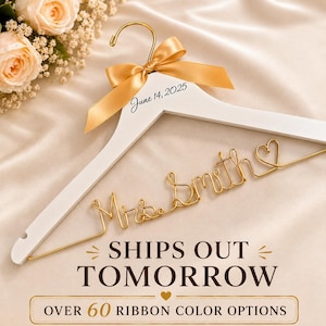 May include: A white wooden clothes hanger featuring a gold wire script reading Mrs. Smith with a heart at the end. The hanger includes a gold satin ribbon bow tied at the top hook and the printed text June 14, 2025 on the wooden frame.