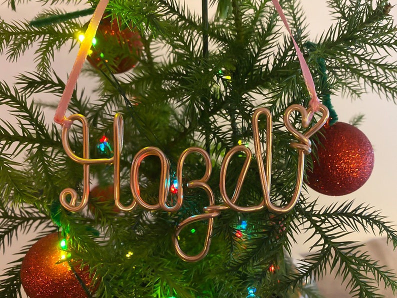 May include: A copper wire Christmas ornament spelling "Hazel" in cursive script, hanging from a pink ribbon. The ornament is displayed on a Christmas tree with red and gold ornaments and colorful lights.