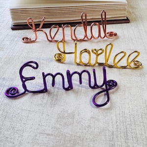May include: Three wire name signs in copper, gold, and purple. The names "Kendal", "Harlee", and "Emmy" are displayed in a cursive font. A book is in the background.