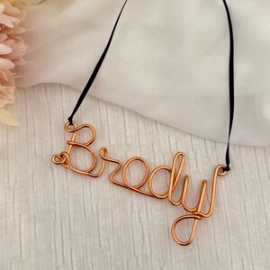 May include: A copper wire name tag spelling "Brody" with a black ribbon for hanging. The name is crafted in a cursive font. The background is a white, textured surface with a soft, floral accent.