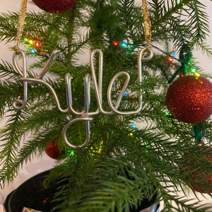 May include: A Christmas tree with red glitter ornaments and a silver wire ornament spelling "Rule!". The tree is decorated with colorful lights and is set against a white background. The ornament is hanging from a gold string.