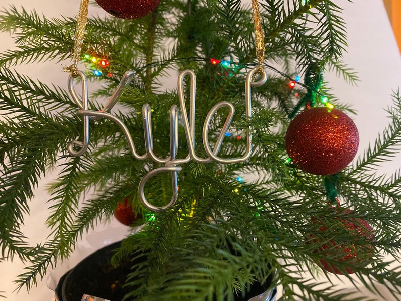 May include: A Christmas tree with red glitter ornaments and a silver wire ornament spelling "Rule!". The tree is decorated with colorful lights and is set against a white background. The ornament is hanging from a gold string.