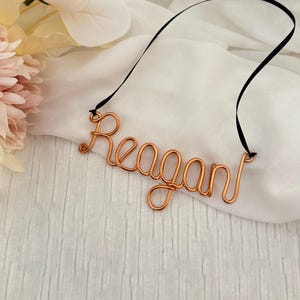 May include: Copper wire name "Reagan" with a black ribbon, displayed on a white fabric background. The name is crafted in a cursive font. Soft pink and white flowers are in the upper left corner.