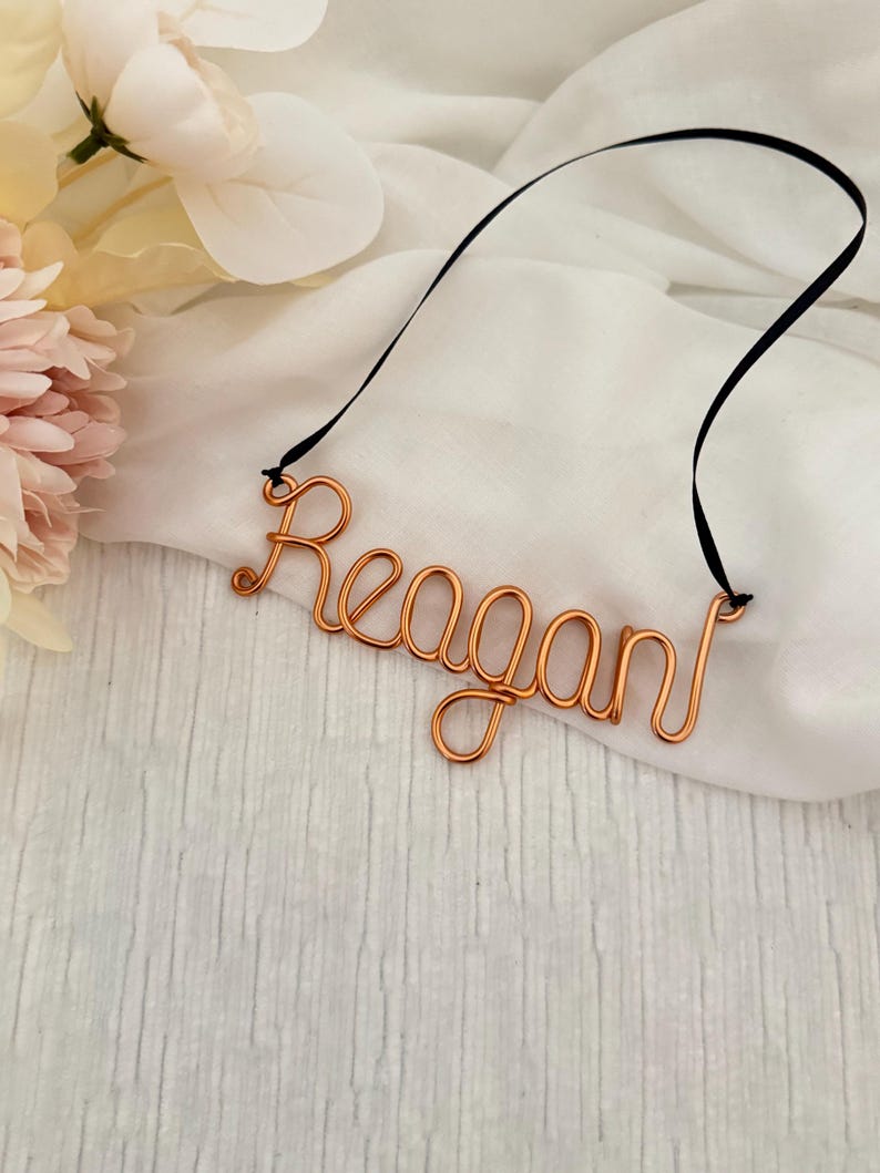 May include: Copper wire name "Reagan" with a black ribbon, displayed on a white fabric background. The name is crafted in a cursive font. Soft pink and white flowers are in the upper left corner.