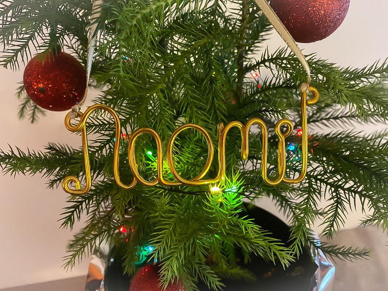 May include: A gold wire ornament spelling "Naomi" hangs from a Christmas tree. The ornament is suspended by a cream-colored ribbon. Red glittery ornaments and colorful lights decorate the evergreen branches.