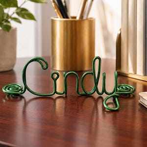 May include: A custom name sign crafted from a single continuous length of metallic green wire, bent into the cursive script spelling the name Cindy. The wire features decorative spiral loops at both the beginning and the end of the name.