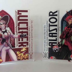 Puede incluir: Dos soportes acrílicos de personajes. Uno presenta a Lucifer con un traje blanco con el texto "LUCIFER". El otro presenta a Alastor con un traje rojo con el texto "ALASTOR". Cada soporte tiene arte de personajes y citas.