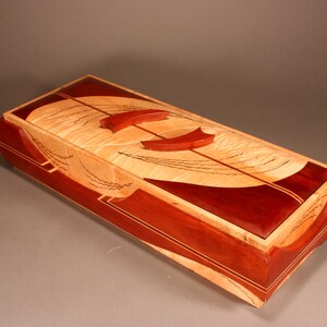 May include: Rectangular wooden box with a light-coloured top featuring a dark red, semi-circular design and two red, curved accents. The box has a dark red border and is likely used for storage.