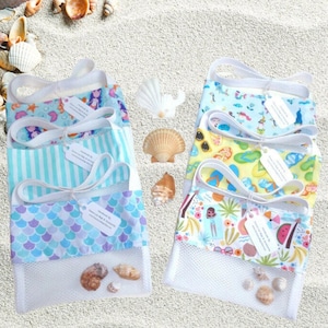 May include: Several beach-themed mesh bags with colorful fabric tops and white handles are displayed on a sandy surface. The bags feature various patterns, including mermaids, stripes, and tropical designs. Seashells are scattered around the bags.
