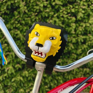 May include: A yellow and black lion head bicycle accessory attached to the handlebars of a red bicycle. The lion has a white muzzle and red mouth. Rainbow streamers are attached to the handlebars.