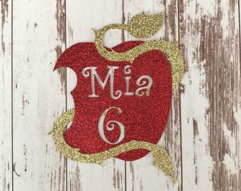 Pode incluir: Maçã vermelha com glitter com um caule e folhas de glitter dourado. A maçã tem uma mordida e o nome "Mia" e o número "6" estão escritos em branco na maçã.