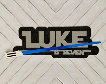 May include: Black and white felt banner with the name "LUKE" in silver letters and the text "IS SEVEN" below. A blue lightsaber toy is placed diagonally across the banner.