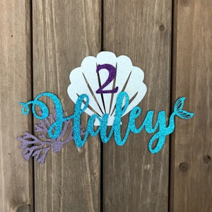 May include: A turquoise glitter mermaid name topper with a white glitter seashell and a purple glitter number 2. The name "Hadley" is written in a cursive font.