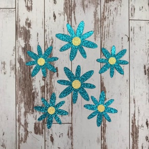 May include: Six glittery blue daisy cutouts with yellow centers are arranged on a light wood-grain surface. The flowers have multiple petals and a sparkling texture, perfect for craft projects or decorations.