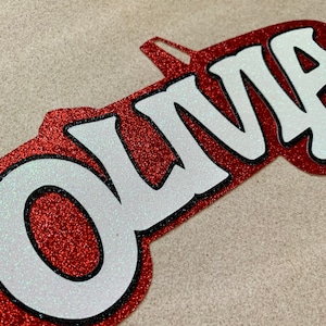 May include: Red glitter name cutout with the name "OLIVIA" in white letters with black outlines.