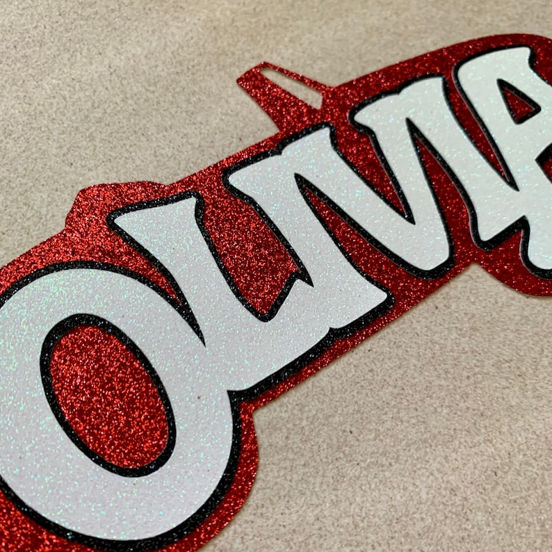 May include: Red glitter name cutout with the name "OLIVIA" in white letters with black outlines.
