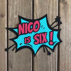 May include: A blue and black comic book style speech bubble with white polka dots. The text "NICO IS SIX!" is in red letters.