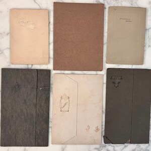 May include: An overhead shot of six vintage paper items. The items include a light tan paper with handwritten text, a brown paper, a light tan paper with the text "Photomaton Studios", and three dark gray paper items with decorative elements.