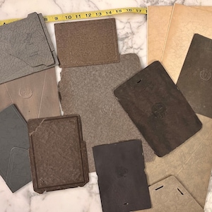 May include: A collection of vintage paper ephemera in various shades of gray, brown, and beige. The items include folders, envelopes, and booklets, some with embossed designs and text. A measuring tape is visible.