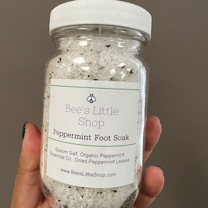 Puede incluir: Un frasco de vidrio lleno de sal de Epsom blanca, aceite esencial de menta orgánica y hojas de menta secas. El frasco tiene una tapa blanca y una etiqueta que dice "Bee's Little Shop Peppermint Foot Soak" e incluye la dirección del sitio web "www.BeesLittleShop.com".