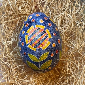 May include: A hand-painted, decorative egg with a blue background and a floral design. The flower has yellow, red, and orange petals with a striped center. The egg is decorated with red and yellow dots and green leaves.