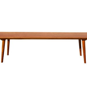 May include: A long, rectangular wooden coffee table with a light brown finish. The table has four slender legs that taper towards the bottom.