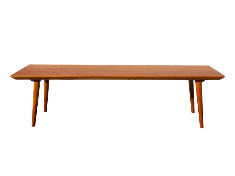 May include: A long, rectangular wooden coffee table with a light brown finish. The table has four slender legs that taper towards the bottom.