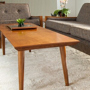 May include: A mid-century modern coffee table with a light wood finish and tapered legs. The table is positioned in front of a gray sofa and a gray armchair.