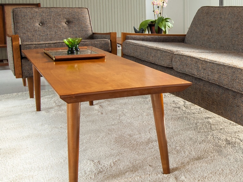 May include: A mid-century modern coffee table with a light wood finish and tapered legs. The table is positioned in front of a gray sofa and a gray armchair.