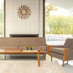 May include: A mid-century modern living room with a brown sofa, a matching armchair, and a wooden coffee table. The room has a white tiled fireplace and large windows overlooking a backyard with a swimming pool.