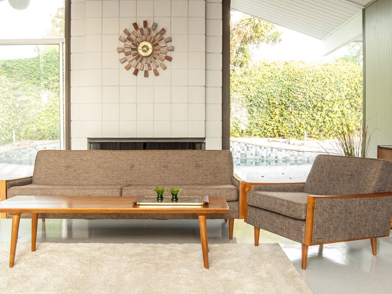 May include: A mid-century modern living room with a brown sofa, a matching armchair, and a wooden coffee table. The room has a white tiled fireplace and large windows overlooking a backyard with a swimming pool.