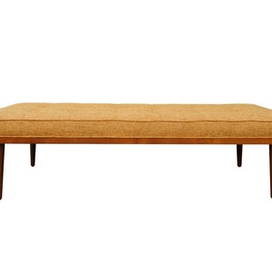 May include: A long, rectangular, brown wooden bench with a light brown fabric cushion. The bench has four legs and a simple, mid-century modern design.