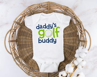 May include: White baby onesie with the text "Daddy's Golf Buddy" in green and blue lettering. A green golf ball with white dimples is in the center of the text.