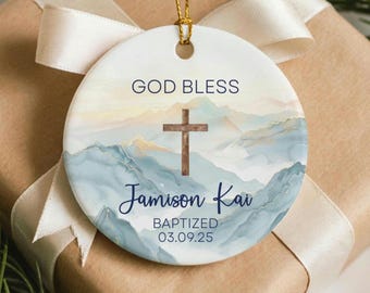 May include: A round ceramic ornament with a gold string for hanging. The ornament features the words "GOD BLESS" above a wooden cross. Below the cross is the name "Jamison Kai" and the word "BAPTIZED" with a date. The background shows a watercolor mountain scene.