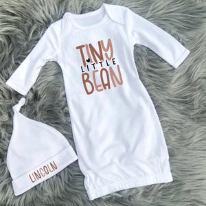 May include: A white baby gown and matching hat set. The gown features the words "TINY LITTLE BEAN" in brown lettering with a small black heart. The hat is embroidered with the name "LINCOLN" in brown. The set is designed for newborns.