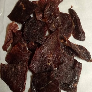 May include: A close-up of a pile of dark brown beef jerky. The jerky is sliced into thin, irregular shapes and has a slightly shiny surface.